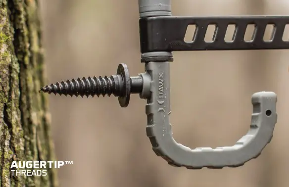 A screw-in tree hanger with a rubber-coated hook is fixed into a tree. The hook is gray with a product logo, conveying durability and practicality.
