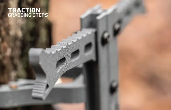 Close-up of a metal tree climbing step with a textured, traction-enhancing design attached to a tree trunk. Text reads "TRACTION GRABBING STEPS." The background is blurred.
