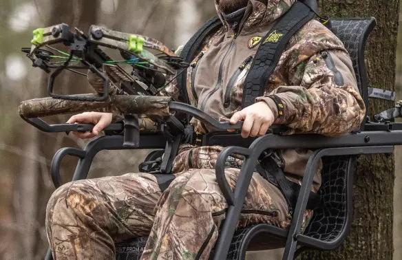 Person in camouflage gear sits in a treestand holding a crossbow, surrounded by a forest setting. The mood is focused and alert.