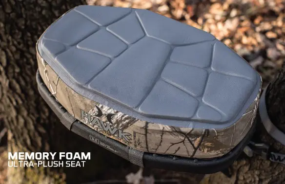 Memory foam hunting seat with a grey, geometric pattern, secured to a tree. The camouflage fabric blends with the forest floor, conveying comfort and durability.
