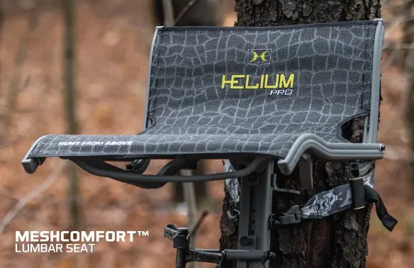 A tree-mounted Helium Pro seat with a gray, patterned mesh design is securely strapped to a tree trunk in a forest setting during autumn.