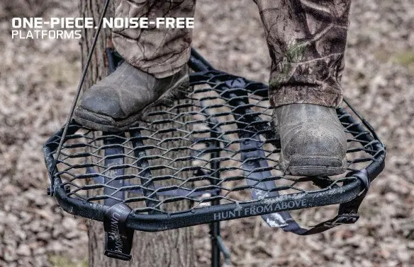 A person stands on a tree platform with sturdy boots. The platform is a black, mesh-like structure, designed for noise-free hunting. Text reads "One-piece, noise-free platforms." Dry leaves cover the ground below, indicating a forest setting.