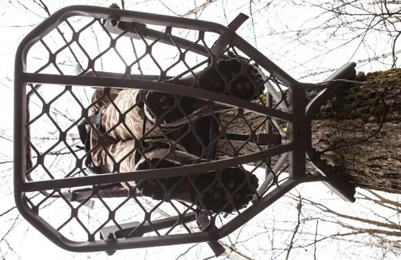 A hunter in camouflage is seated on a metal tree stand, secured to a tree trunk, against a backdrop of bare branches and a cloudy sky.