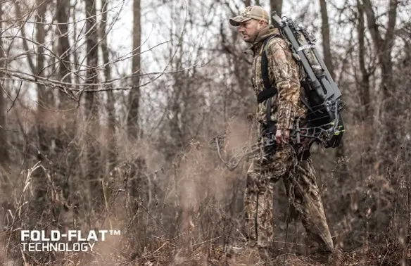 Hunter in camo gear walks through a leafless forest, carrying a crossbow with fold-flat technology. The scene is earthy and focused.