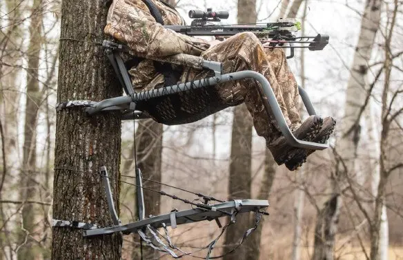 Hunter in camo gear sits in a tree stand, holding a crossbow. The wooded background and wintery trees create a focused, stealthy atmosphere.