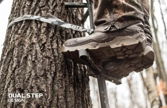 A person in camo pants is climbing a tree ladder, wearing muddy boots. The scene conveys a rugged, outdoorsy vibe. Text reads "Dual Step Design."