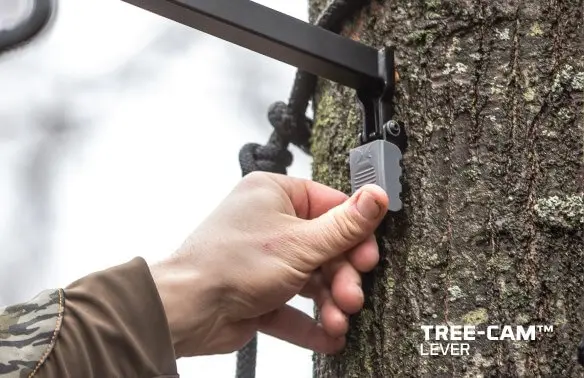A hand adjusts a Tree-Cam lever on a tree, secured with a black rope. The bark texture and camouflage jacket sleeve suggest an outdoor setting.